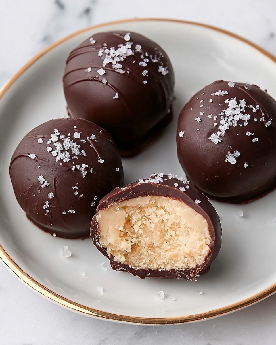 The image shows four round chocolate-covered truffles on a white plate with a thin gold rim, placed on a white marbled surface. Three truffles are whole, their dark chocolate shells smooth and shiny with small flakes of white sea salt sprinkled lightly on top. One truffle is cut in half, revealing its creamy, light beige, slightly crumbly filling inside. The chocolate shell around the cut piece is thin and cracked at the edges, showing the contrast between the dark shell and the pale filling. photo taken with an iphone --ar 4:5 --v 7