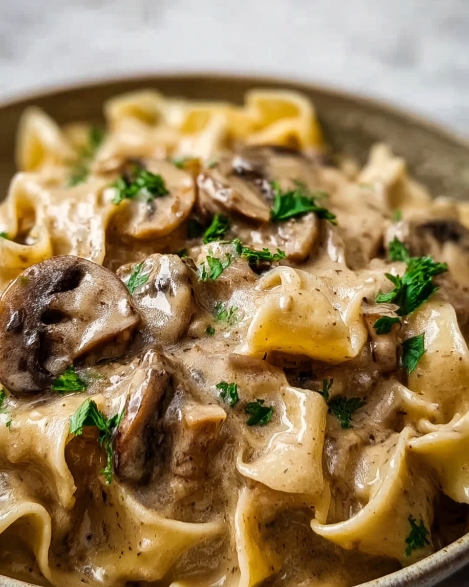 The image shows a close-up of a creamy pasta dish with wide, flat noodles covered in a light brown mushroom sauce. There are several pieces of sliced mushrooms mixed in, all coated with the thick, glossy sauce that looks smooth and rich. Bright green small parsley leaves are sprinkled on top to add a fresh contrast. The dish sits on a white marbled surface, and the focus is tight on the textures of the sauce and pasta. Photo taken with an iphone --ar 4:5 --v 7