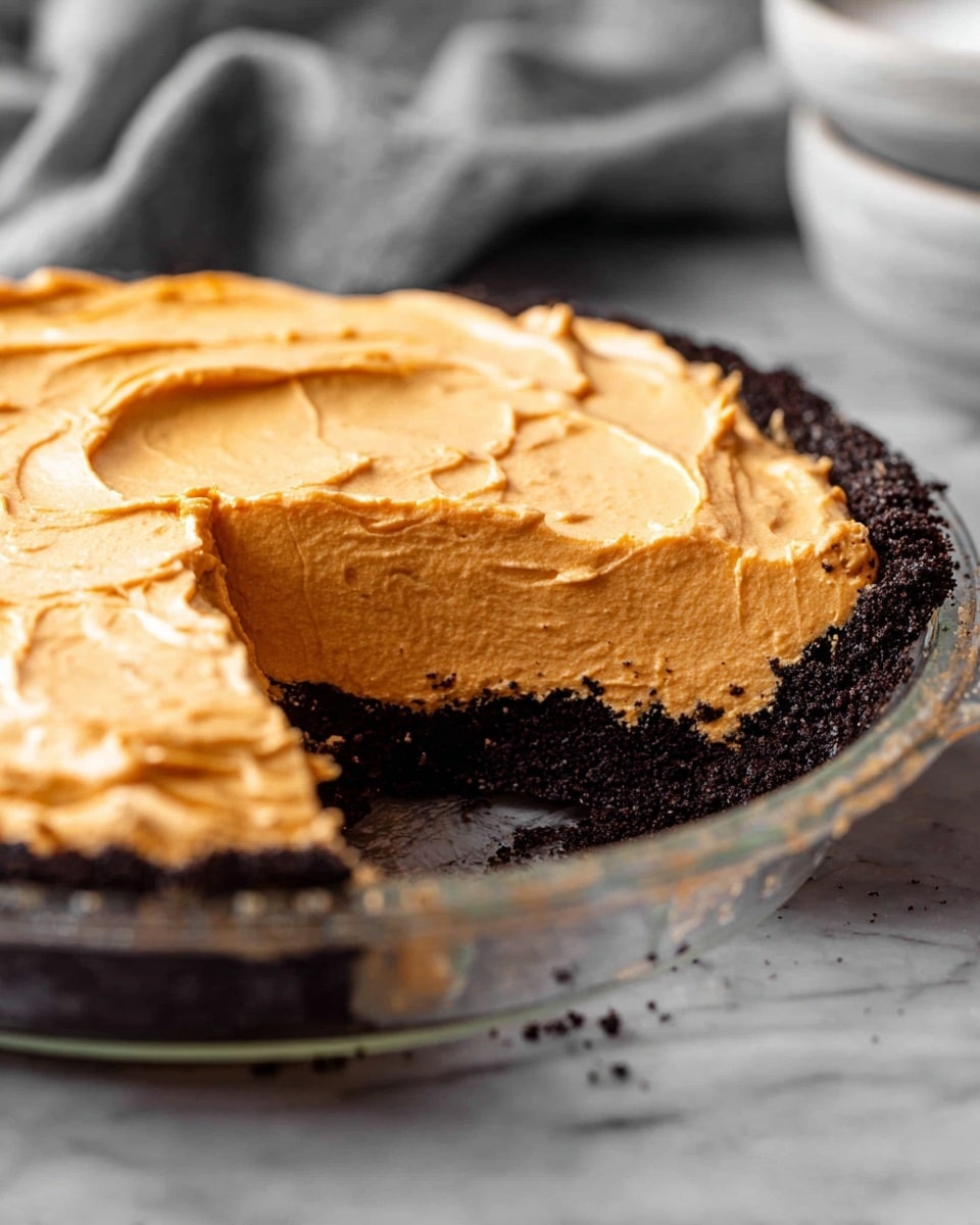A close-up view of a pie with two layers in a clear glass dish on a white marbled surface. The bottom layer is a dark, crumbly chocolate crust, thick and pressed firmly in the dish. The top layer is a tall, light orange, smooth and creamy filling with soft swirls on the top surface. The pie has a slice removed, showing the clean edge between the creamy top and the crumbly bottom. In the background, there is a soft, gray cloth and a blurred white bowl. photo taken with an iphone --ar 4:5 --v 7