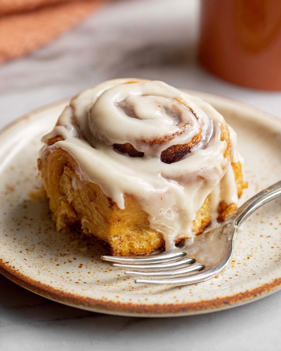 A single cinnamon roll sits on a white plate with small brown speckles, showing about three layers of soft, golden-brown dough with swirls of dark cinnamon filling. The roll is covered evenly with thick, creamy white icing that drips slightly down the sides, creating a smooth and glossy texture. A silver fork rests diagonally on the plate next to the cinnamon roll. The background features a blurred white marbled surface, giving a clean and bright setting. Photo taken with an iphone --ar 4:5 --v 7