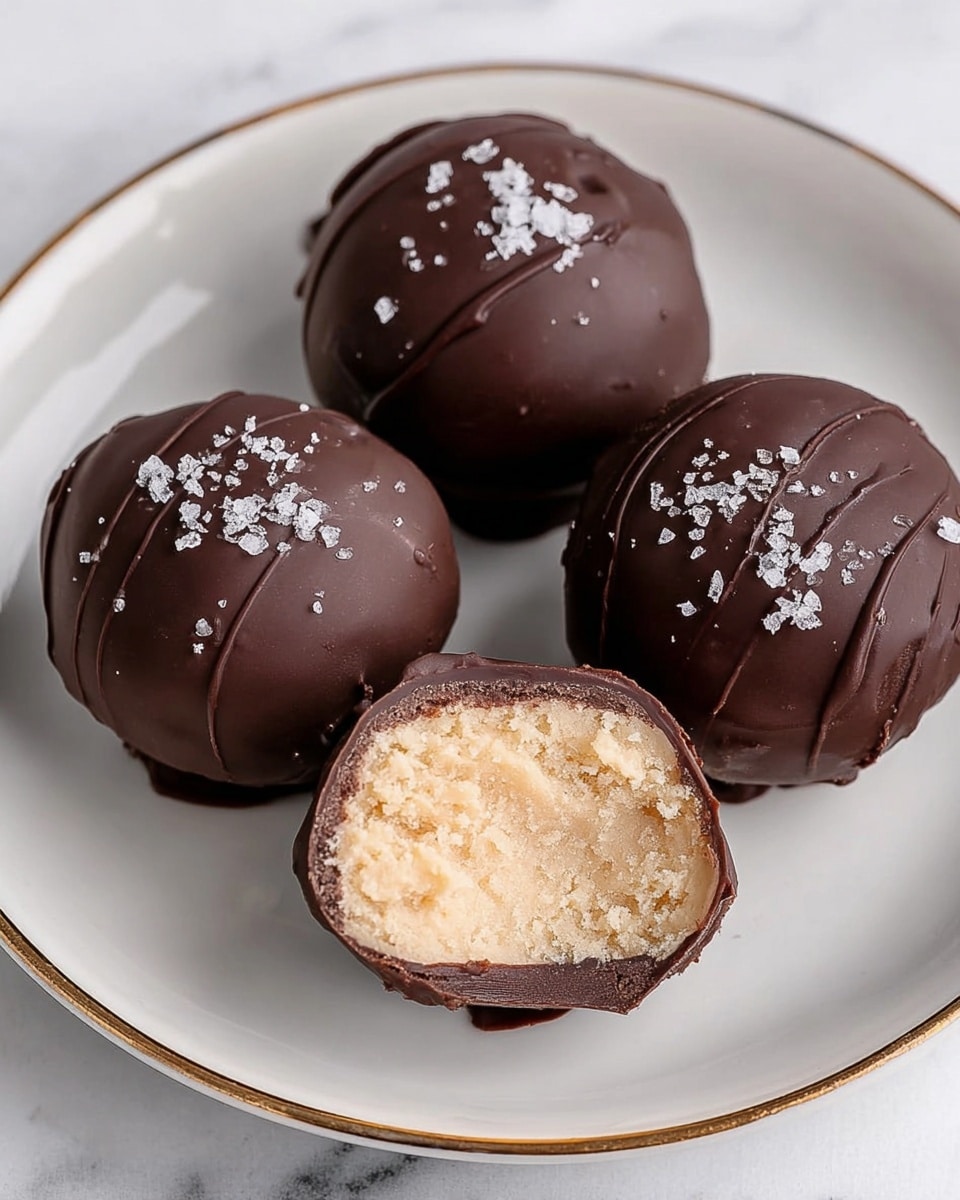 Four round chocolate-covered truffles sit on a white plate with a thin gold rim, placed on a white marbled surface. Three truffles are whole, with smooth, dark chocolate coating sprinkled with coarse white salt on top. One truffle is cut in half, showing a thick, dark chocolate shell surrounding a light, creamy beige filling with a crumbly texture inside. The truffles are arranged close together with the cut one in the front. Photo taken with an iphone --ar 4:5 --v 7